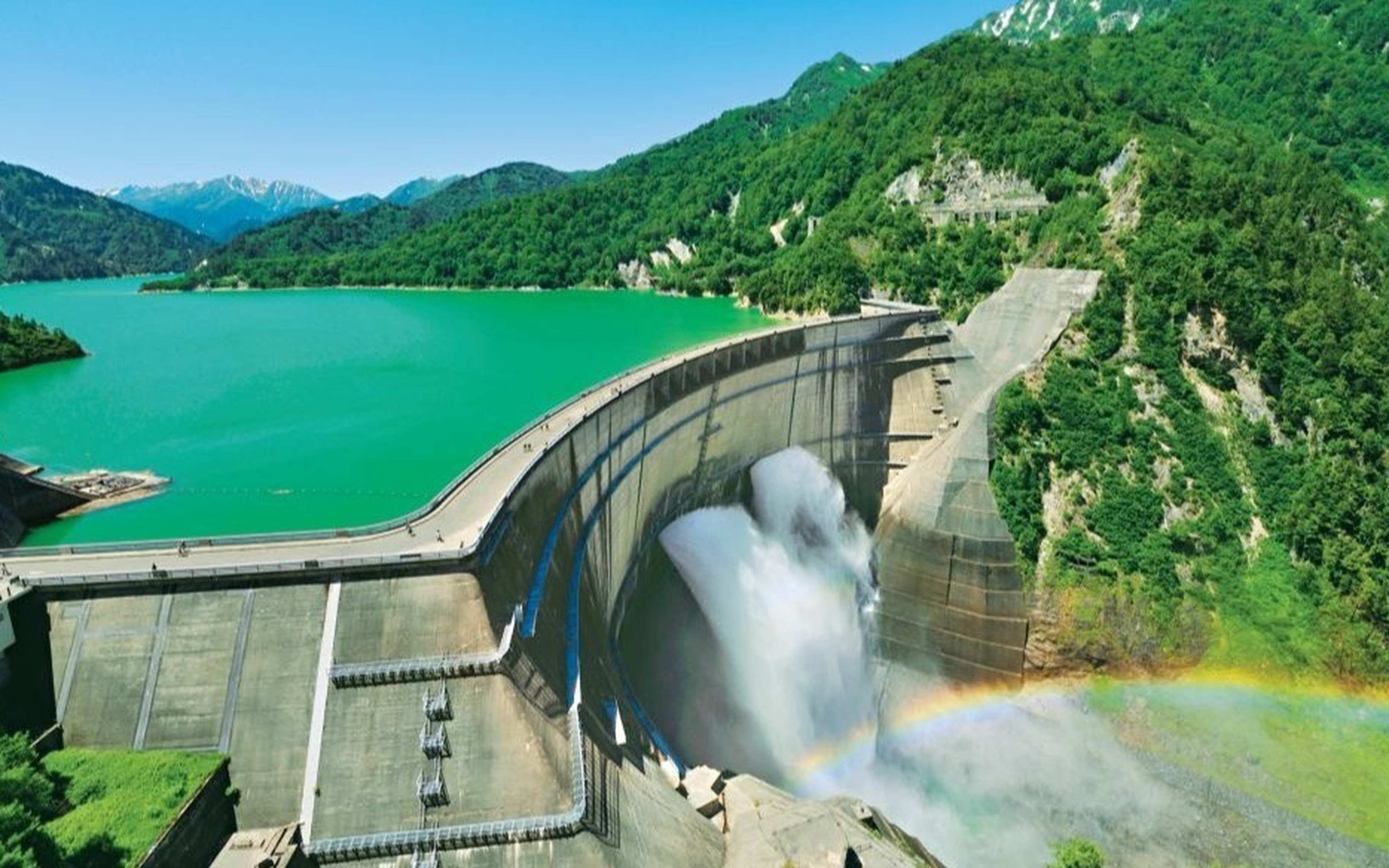 A curved dam releasing water below a green lake, with a rainbow in the spray
