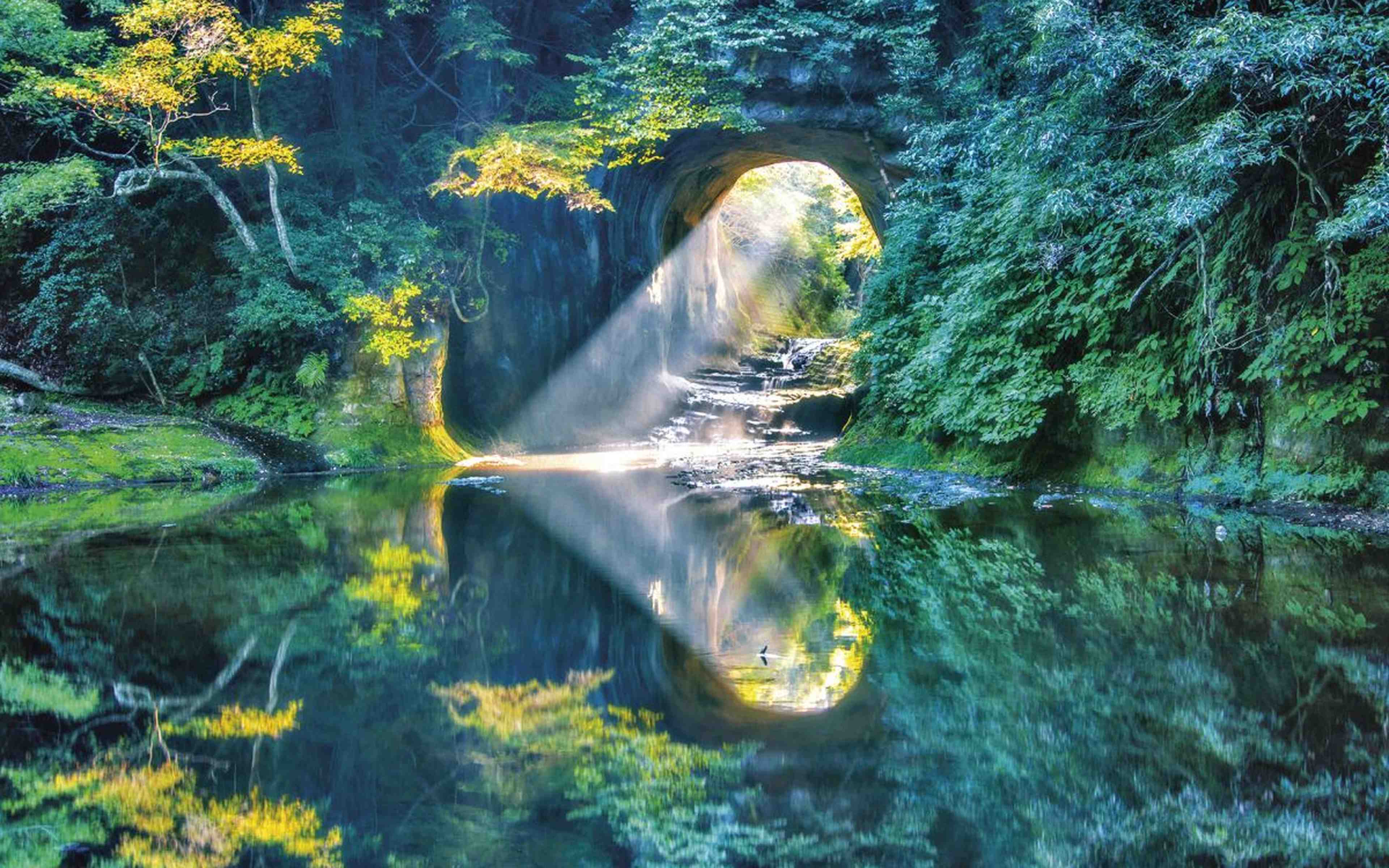 A rock tunnel in a forest shaped like a cave arch, with sunlight streaming through onto a still, reflective water surface